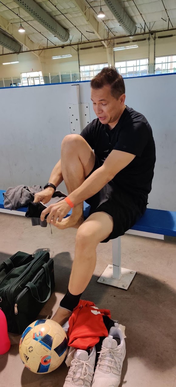 David Gailey sitting on a bench in an indoor sports hall, head down, pulling on a sock, with a soccer ball and kit bag on the floor beside him